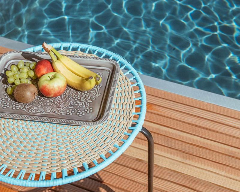  Poolrand mit Gartenmöbel im Vordergrund und Obst auf dem bunten gewebten Tisch 